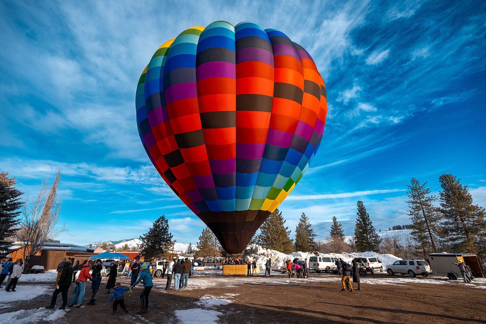 Colorful striped hot-air balloon lifting off over a snowy field with spectators and evergreen trees